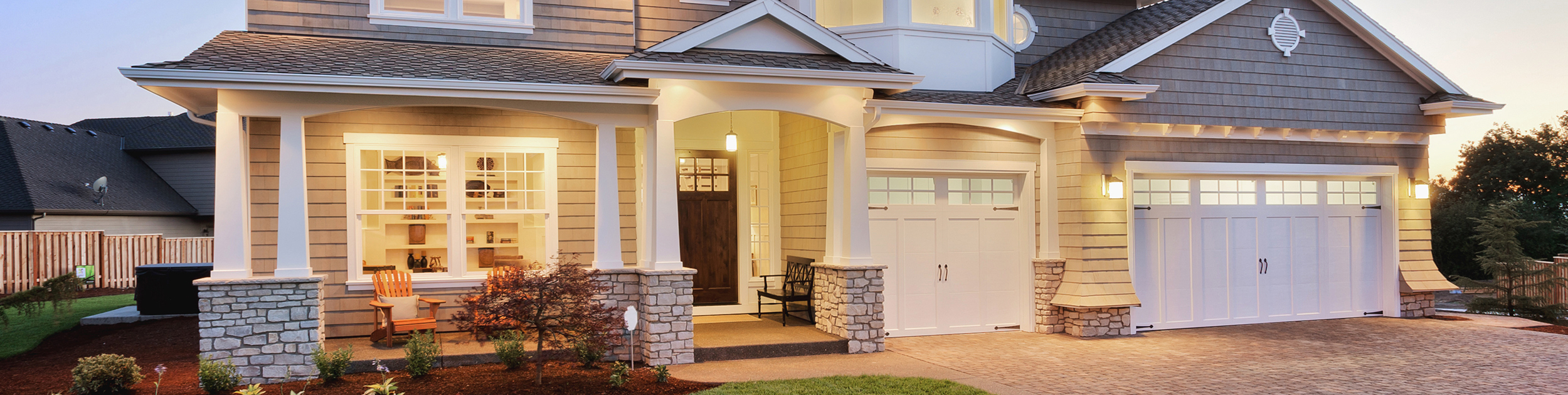 exterior of beautifully lit up home with a stone driveway - gray shingles with white trim and garage doors, stonework and pillars supporting the overhang with two adirondack chairs and a bench on the front porch