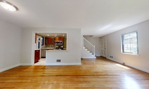 large open empty room with hardwood flooring and light walls with a view of a kitchen through an opening and a staircase on the right side