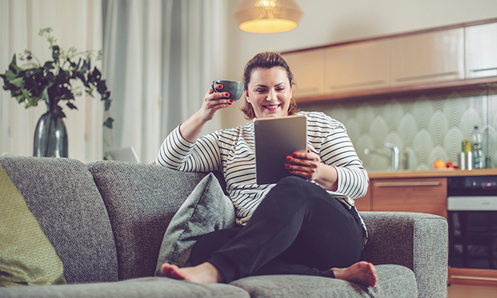 Woman sitting on a couch smiling, holding a cup of coffee in one hand and a tablet in the other.