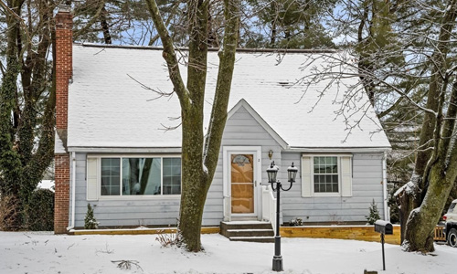 light gray cape style home with snow covered roof, white trim and shutters with a brown door