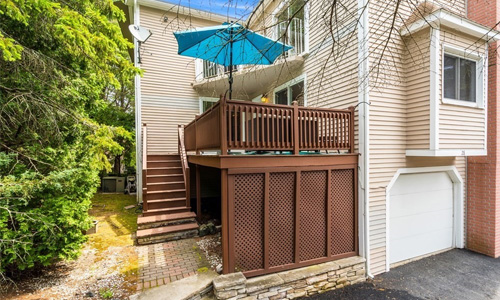 tan residential property showing a beautiful wooden deck with stairs leading up to it and to the right is a white garage door with a bump out window above it