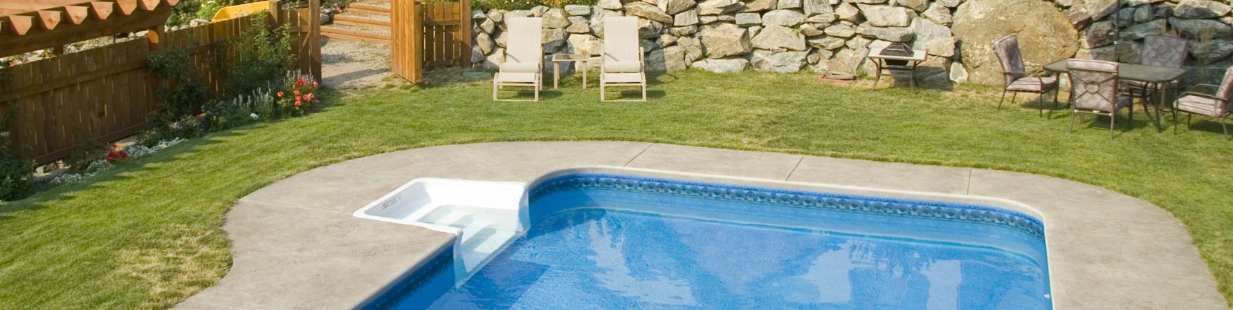 in-ground swimming pool with stone sitting area around it surrounded by grass and a stone wall; some lawn furniture is shown