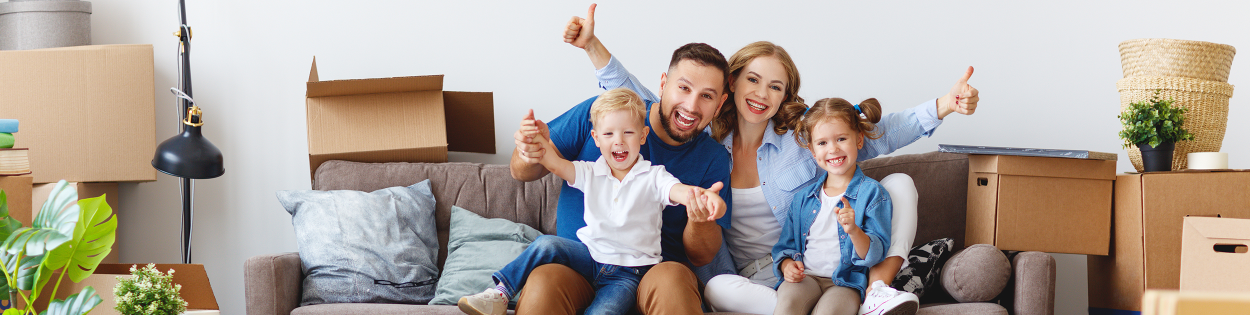 man, woman and two children with big smiles and doing thumbs up while sitting on a couch surrounded by moving boxes