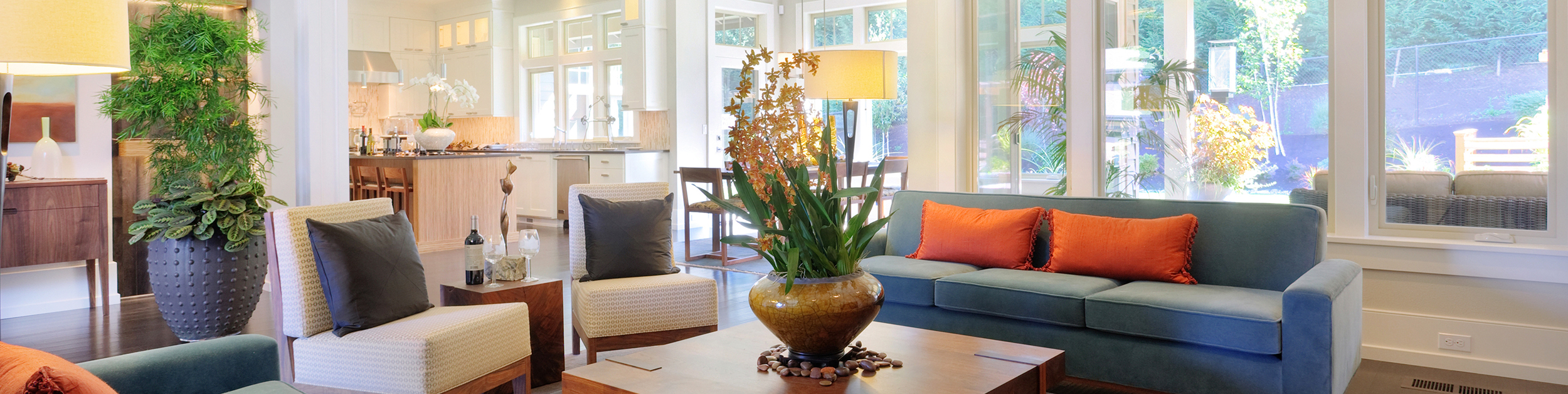 partial view of main living area in a modern open concept home - living room with blue couch and orange pillows as well as white chairs with gray pillows, plants and lamps with the kitchen and dining area in the background all in front of a wall of windows showing the outside