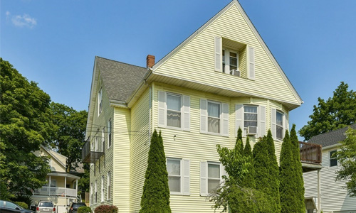 three story yellow house with large peaks on the upper level, white shutters and tall trees out front