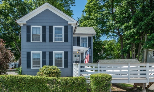49 Hastings Street Marlborough, MA 01752 Blue Colonial style home with white trim, black shutters, covered entranceway with white door, an American flag, and a white fenced in deck shown to the right