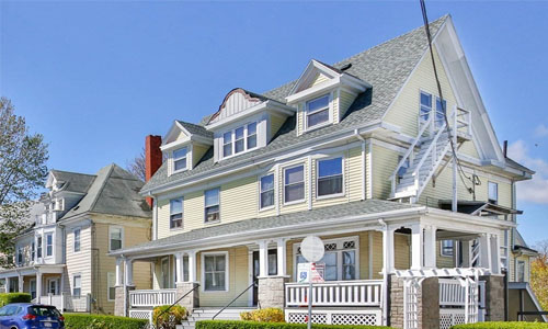 Three family home in Gloucester MA - exterior of property shown - light yellow with farmers porch out front, three dormers and a fire escape shown on the right of the house