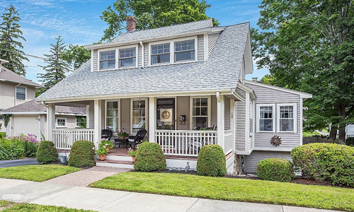Light gray colonial style home with white trim, farmer's porch, well maintained hedges and flower pots out front - surrounded by bare trees and a blue sky